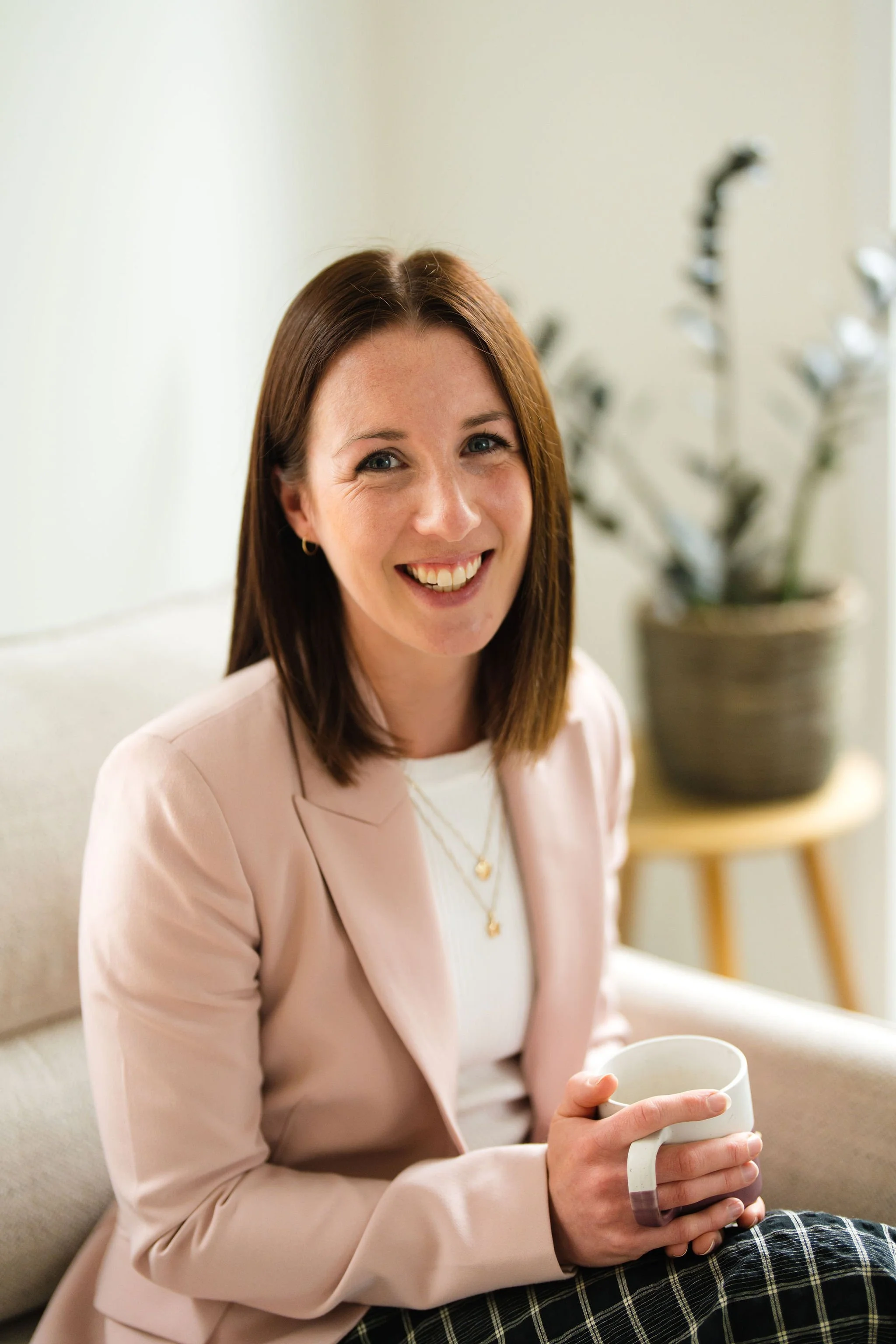 A smiling woman with shoulder-length brown hair, wearing a light pink blazer, white shirt, and gold jewelry, sitting on a beige sofa holding a white mug, with a potted plant in the background.