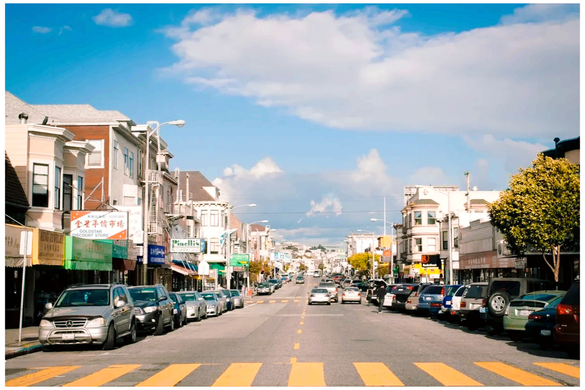 A city street scene with parked cars on both sides, storefronts along the sidewalks, and a large cloud-filled sky overhead.