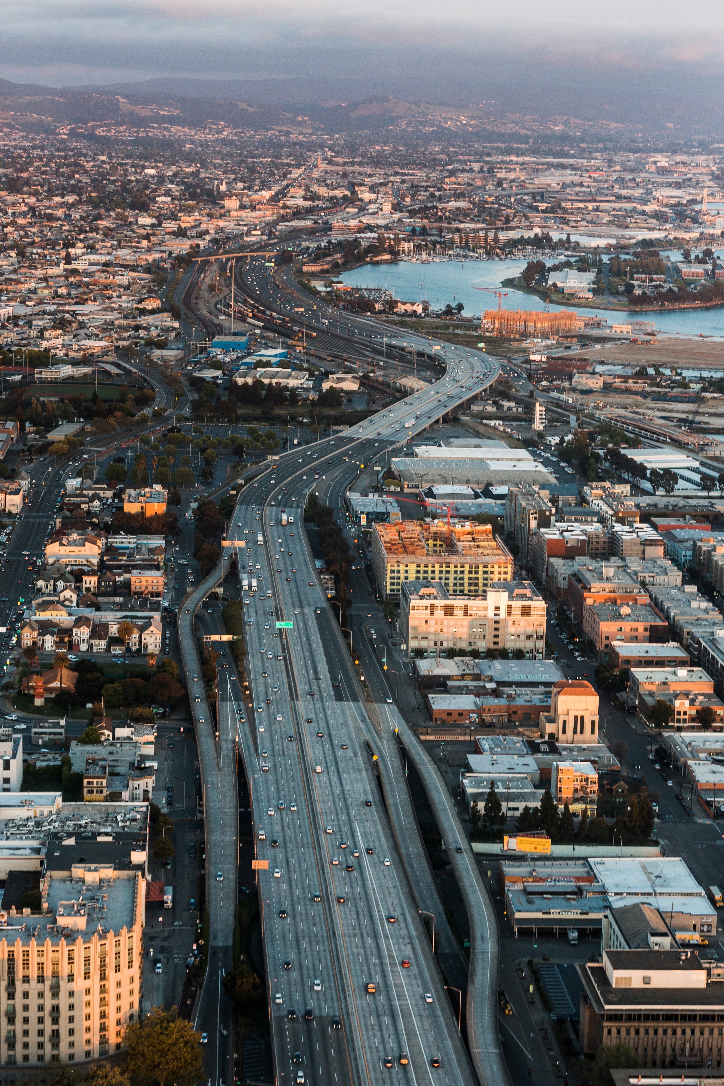 Aerial view of a city with a winding highway, various buildings, and a large body of water, during sunset.