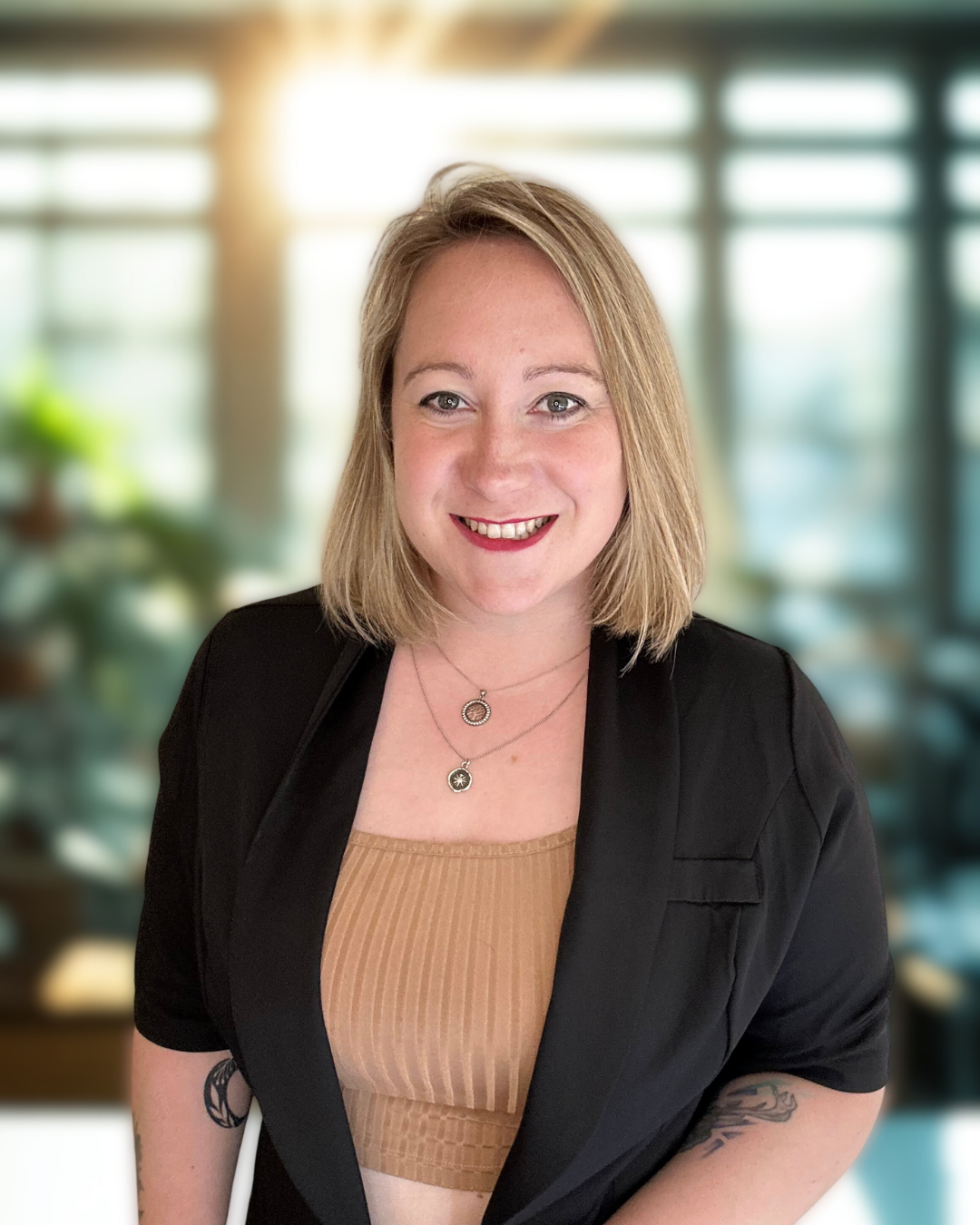 A smiling woman with shoulder-length blonde hair, wearing a beige top and a black blazer, standing in a bright, modern office with large windows and blurred background.