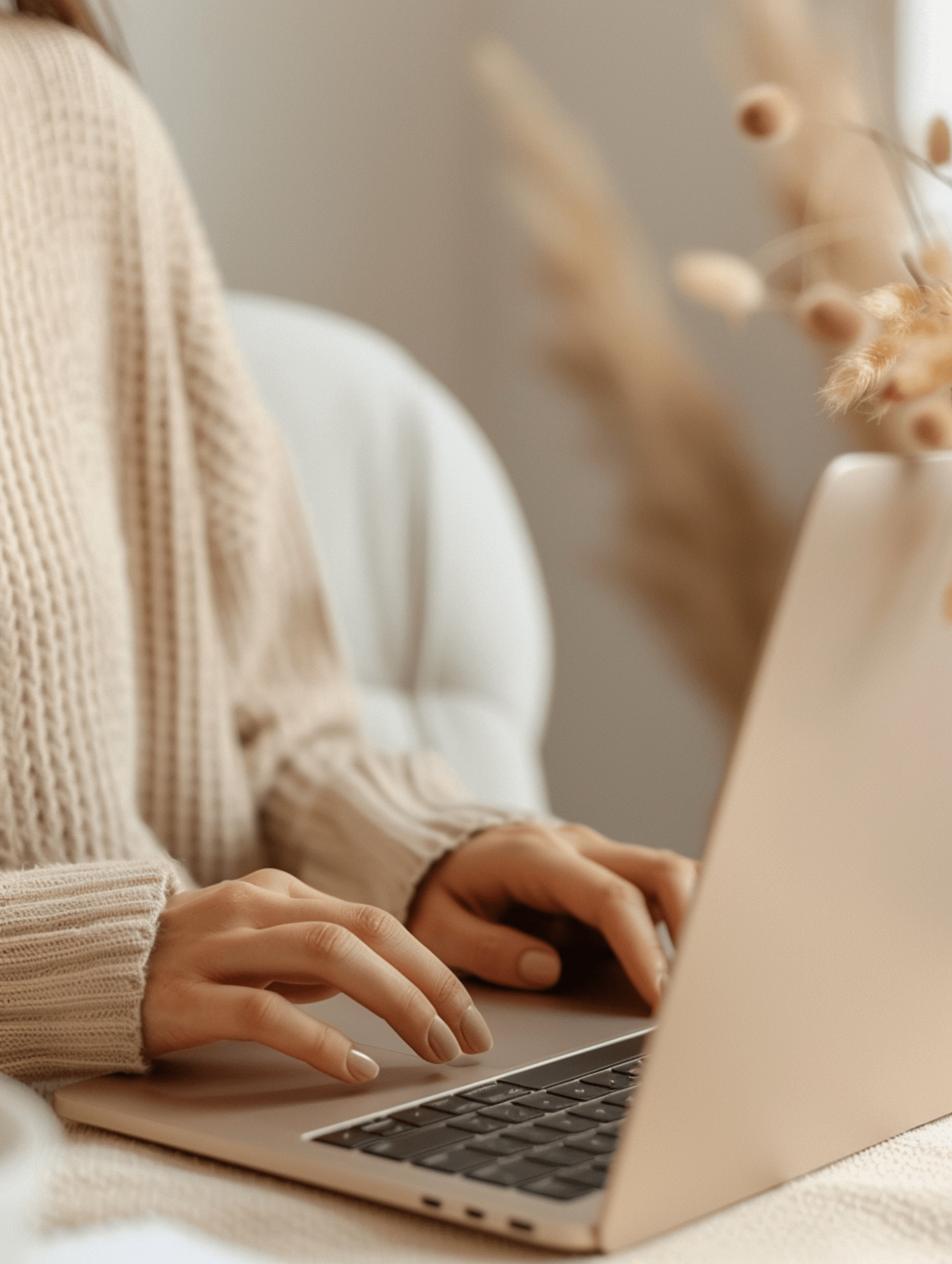 Person wearing a beige knit sweater typing on a laptop with a white chair and beige decorative elements in the background.