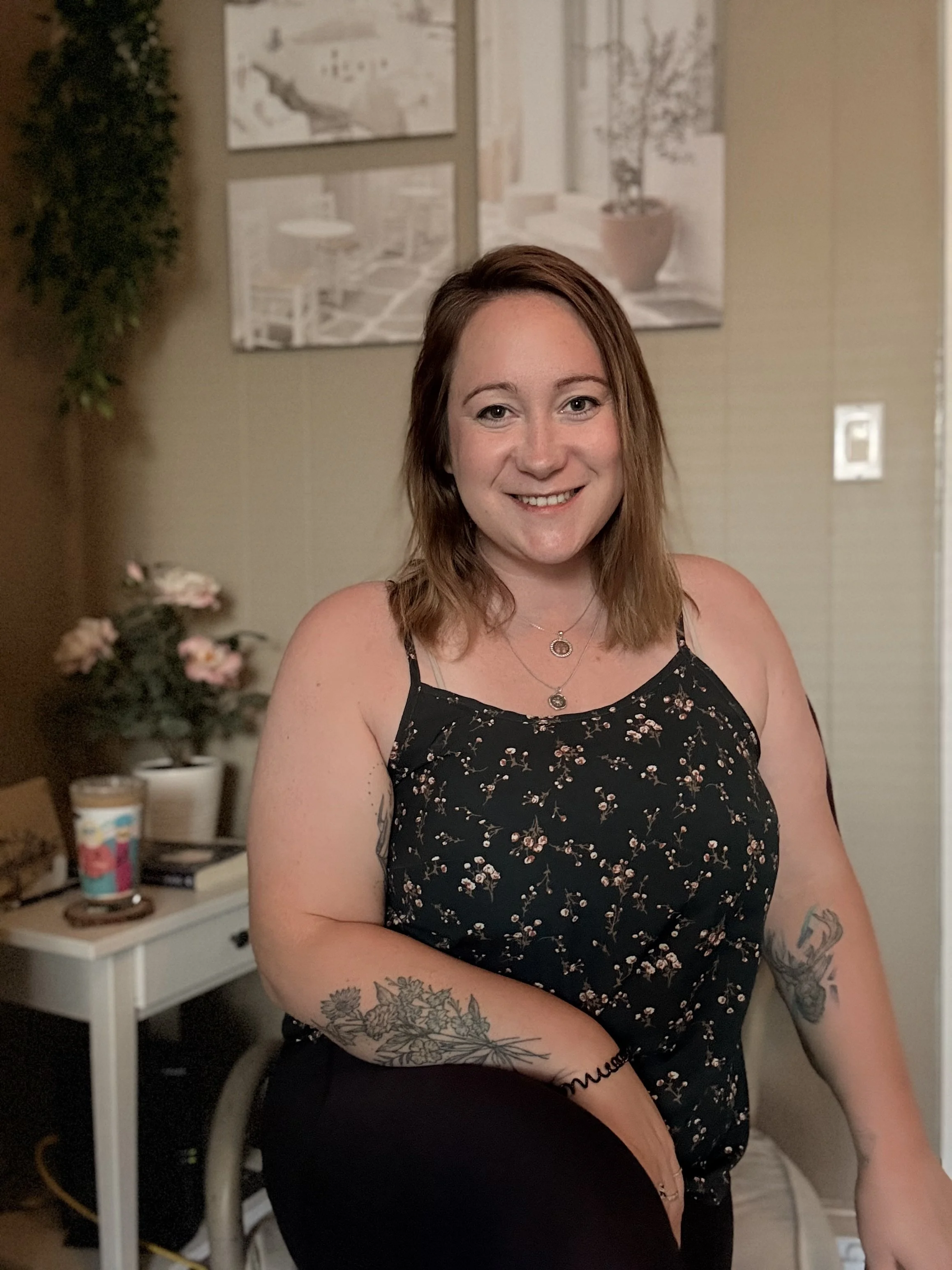 A young woman with shoulder-length red hair and tattoos on her arms, smiling and sitting in a living room. She is wearing a black floral sleeveless top and layered necklaces. Behind her, there is a small white table with a potted plant, a coffee cup, and some papers, along with framed artwork on the wall.