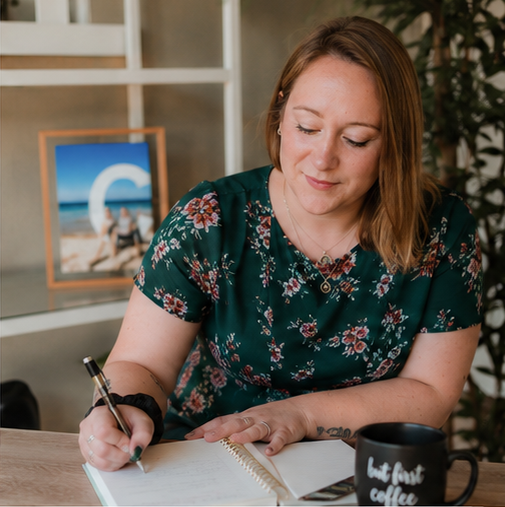Woman with light brown hair wearing a floral green shirt writing in a notebook at a desk, with a framed photograph of a beach scene and a black coffee mug nearby.