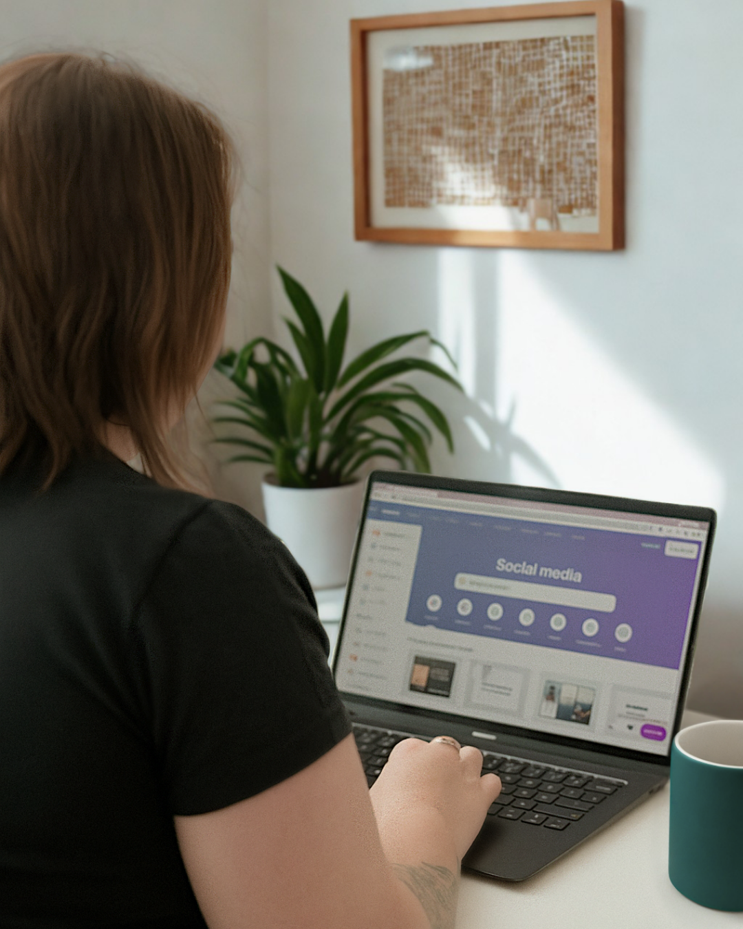 Person with brown hair using a laptop to browse social media setup on a white desk with a teal cup and a potted plant, wall art in the background.