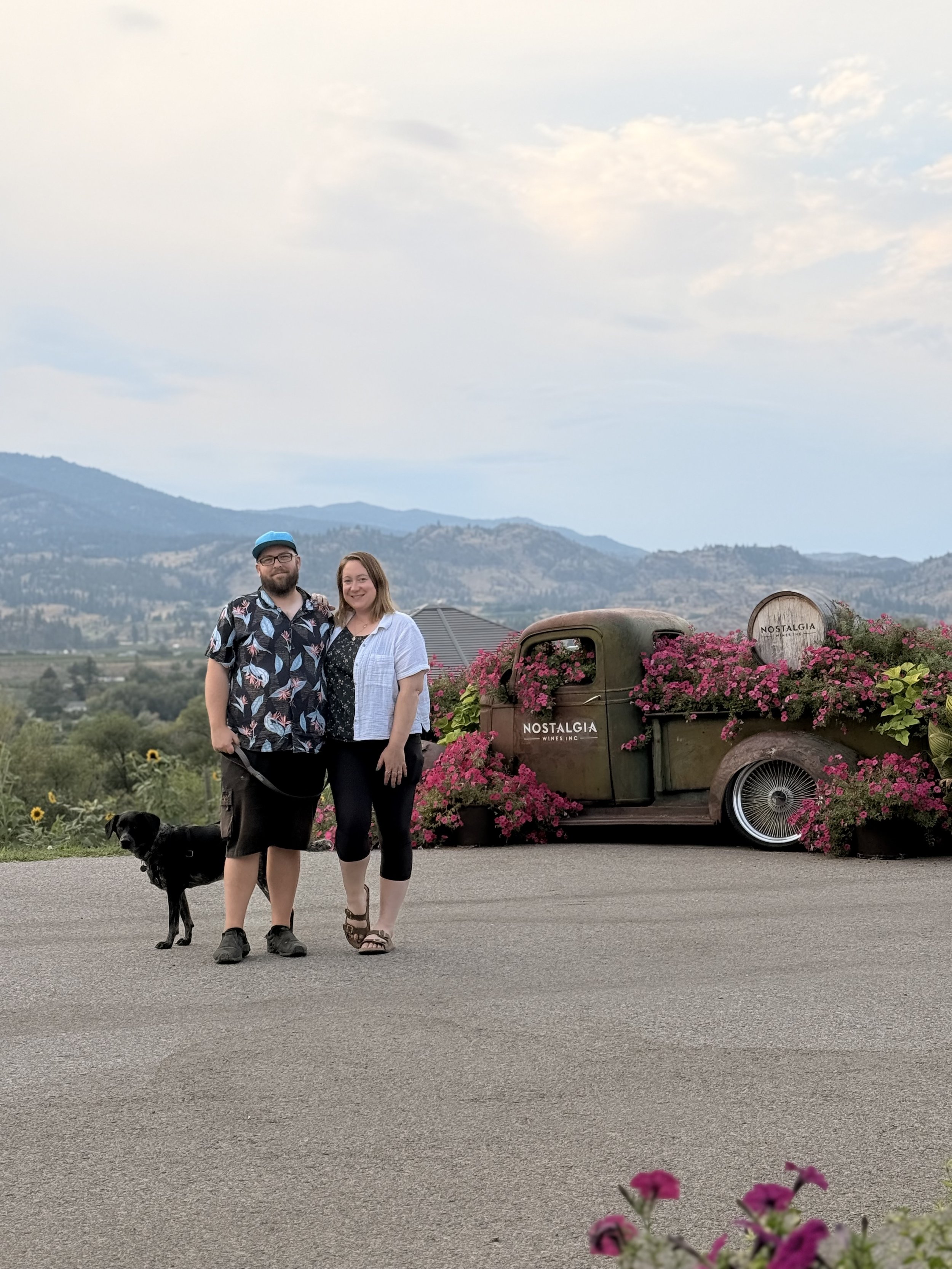 A couple and a black dog standing on a paved surface outdoors near a vintage truck with pink flowers, with mountains and a cloudy sky in the background.