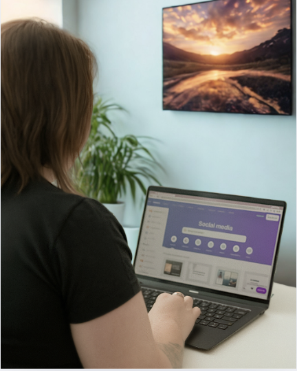 Person using a laptop with social media dashboard open, sitting at a desk with a plant and a wall-mounted TV showing a sunset landscape.