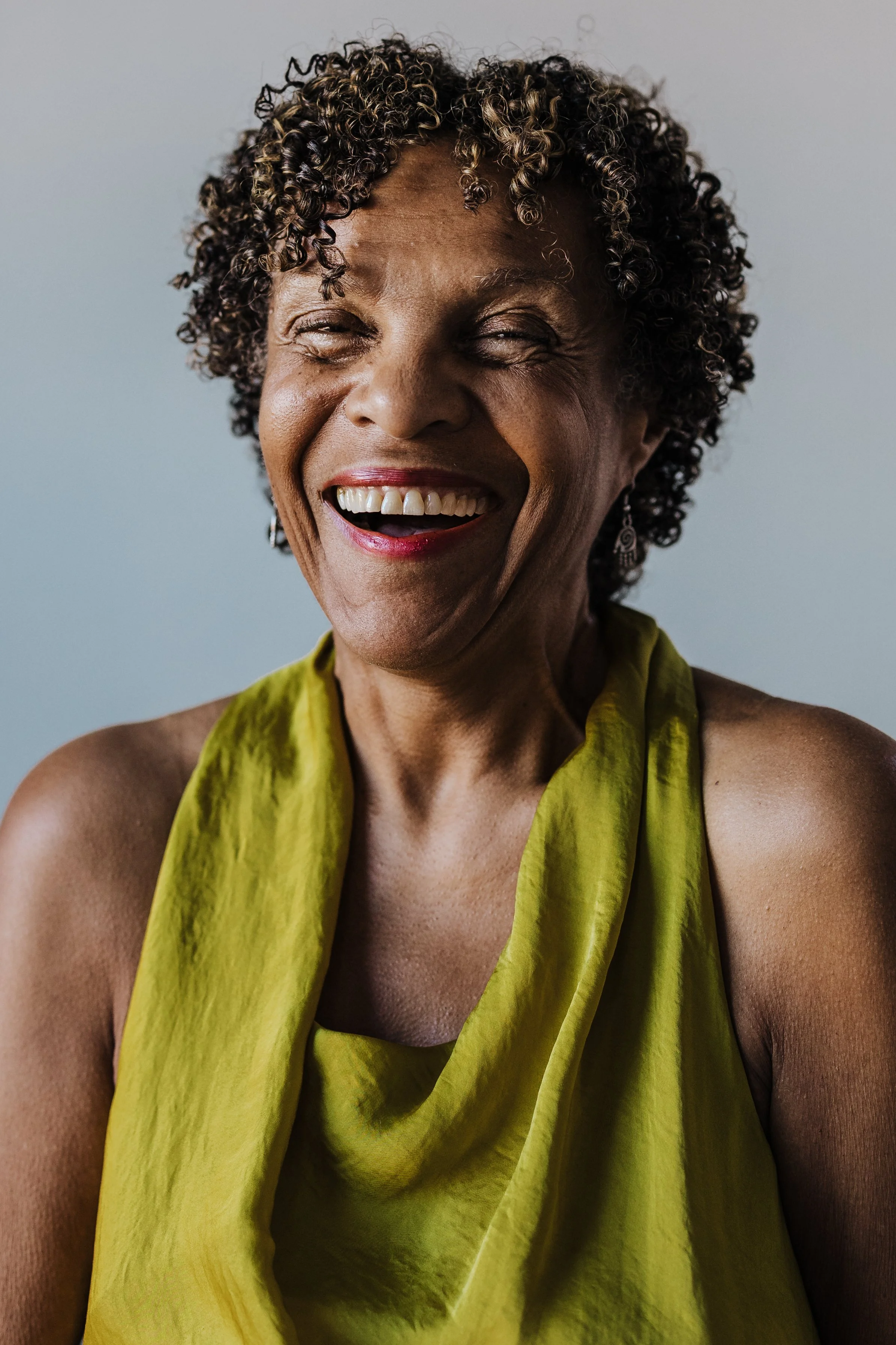 A smiling older woman with short curly hair wearing a sleeveless yellow-green top and earrings.