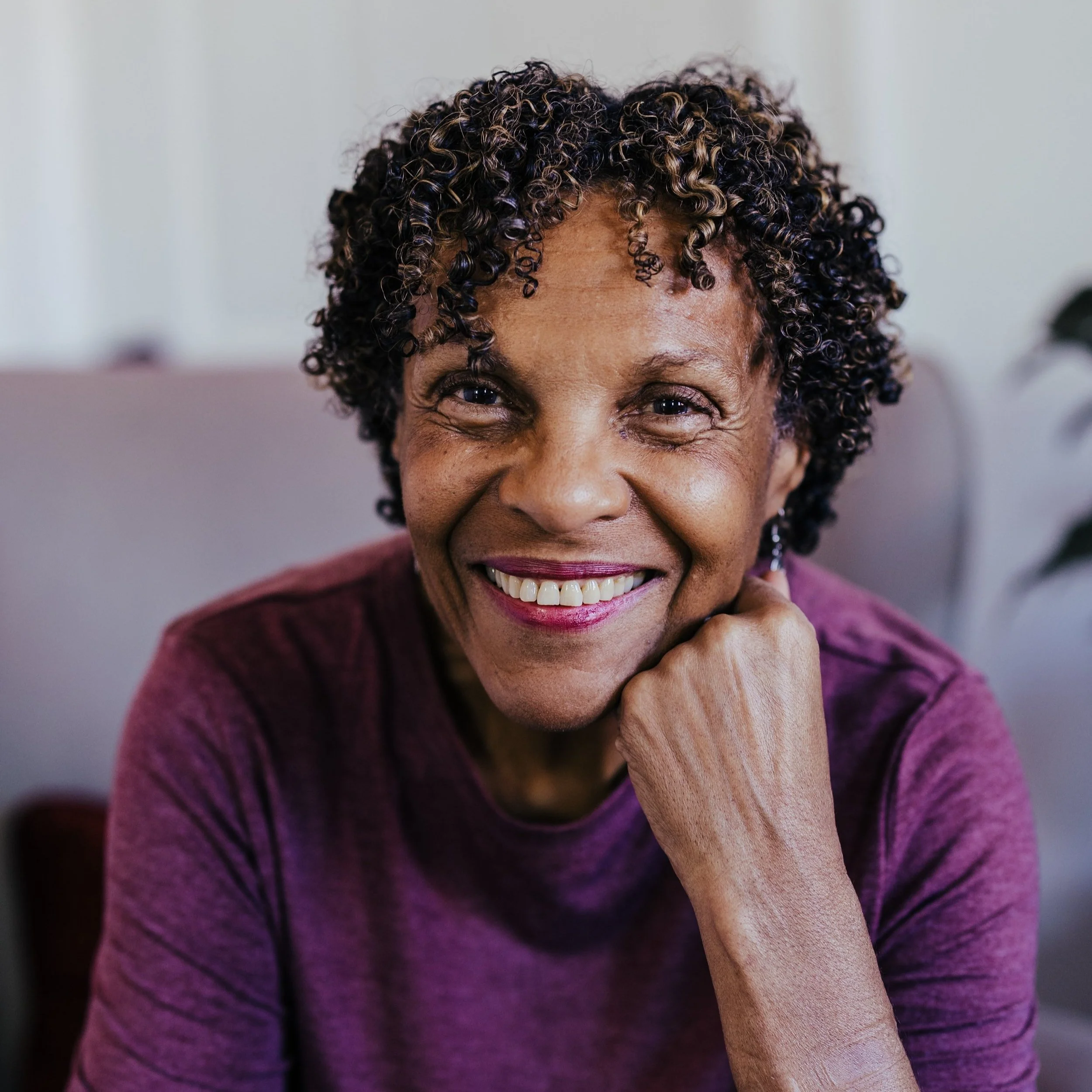 A smiling older woman with short, curly hair, wearing a purple top, sitting indoors with her hand resting on her chin.