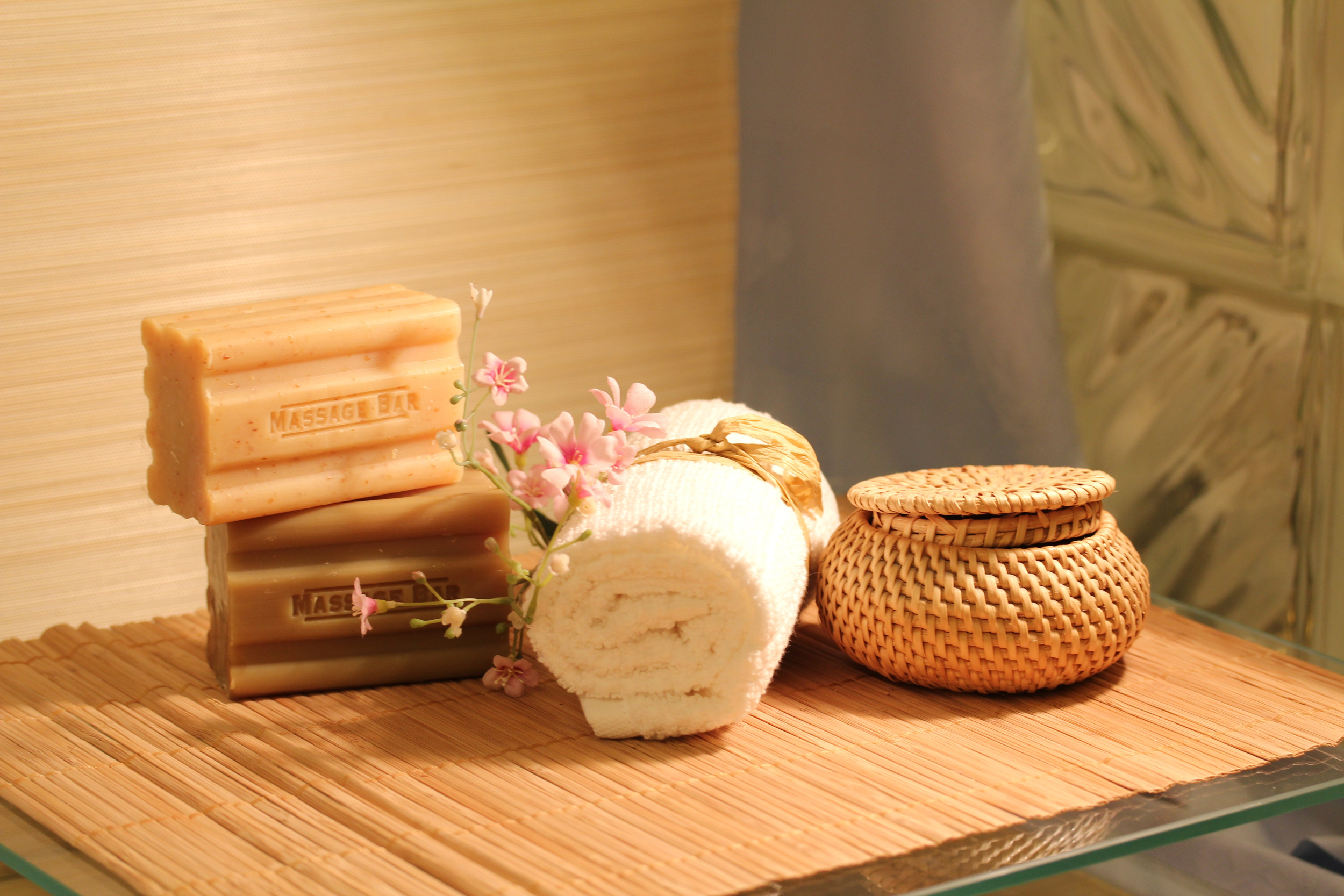 Spa setting with stacked soap bars labeled 'Massage Bar', a rolled white towel, pink flowers, and a small woven basket on a bamboo mat.