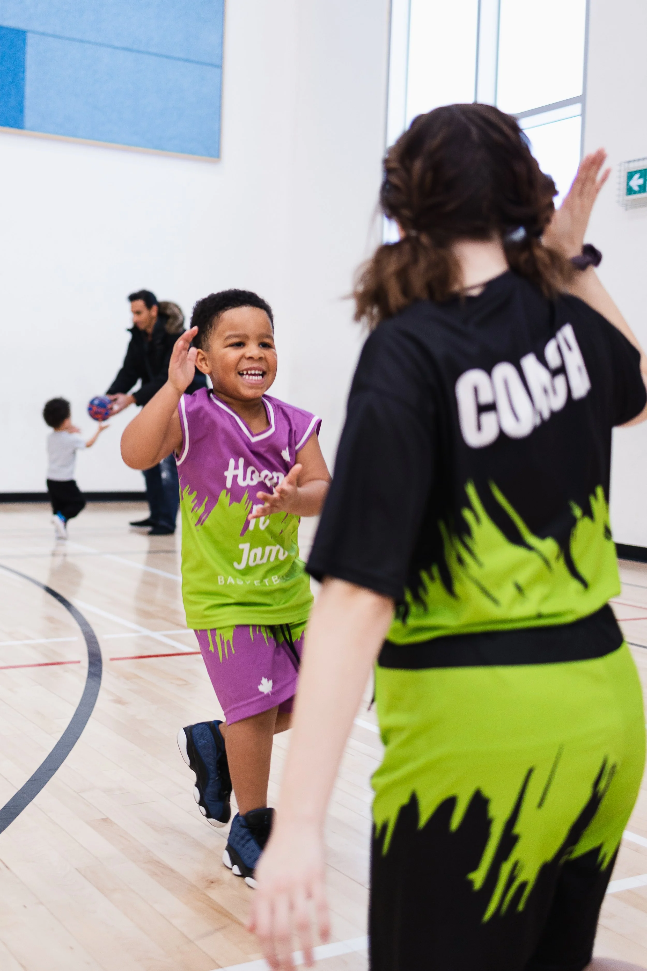 A young girl smiling and receiving a high-five from a sports coach inside a gymnasium, with a man and a young boy playing in the background.