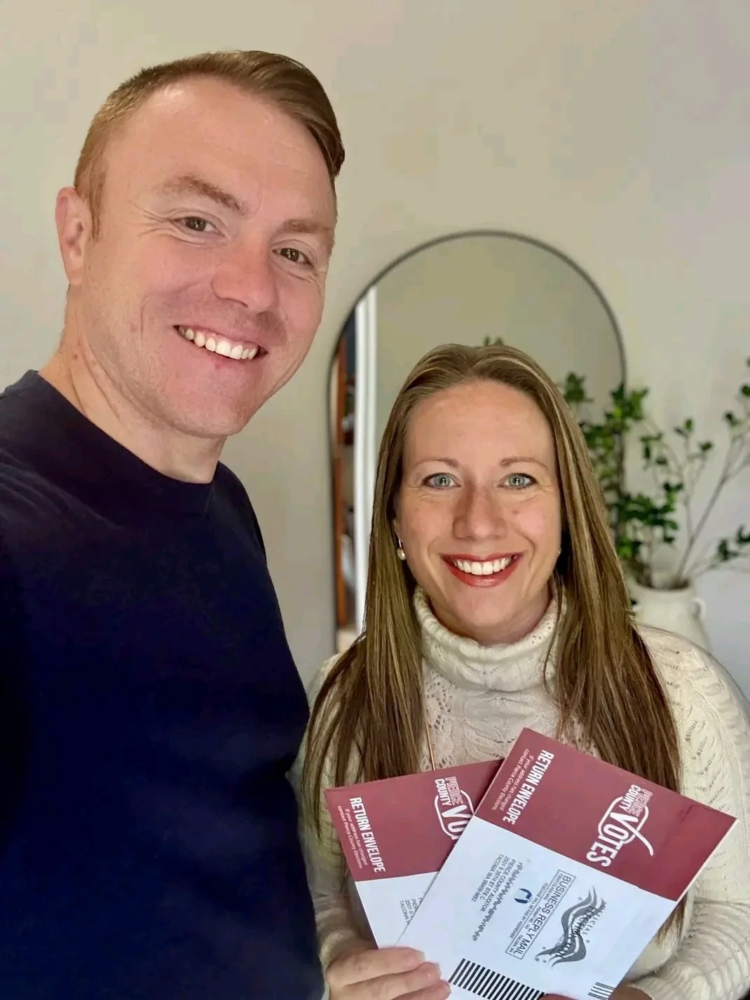 A man and woman smiling indoors, holding ballots with a mirror and plants in the background.