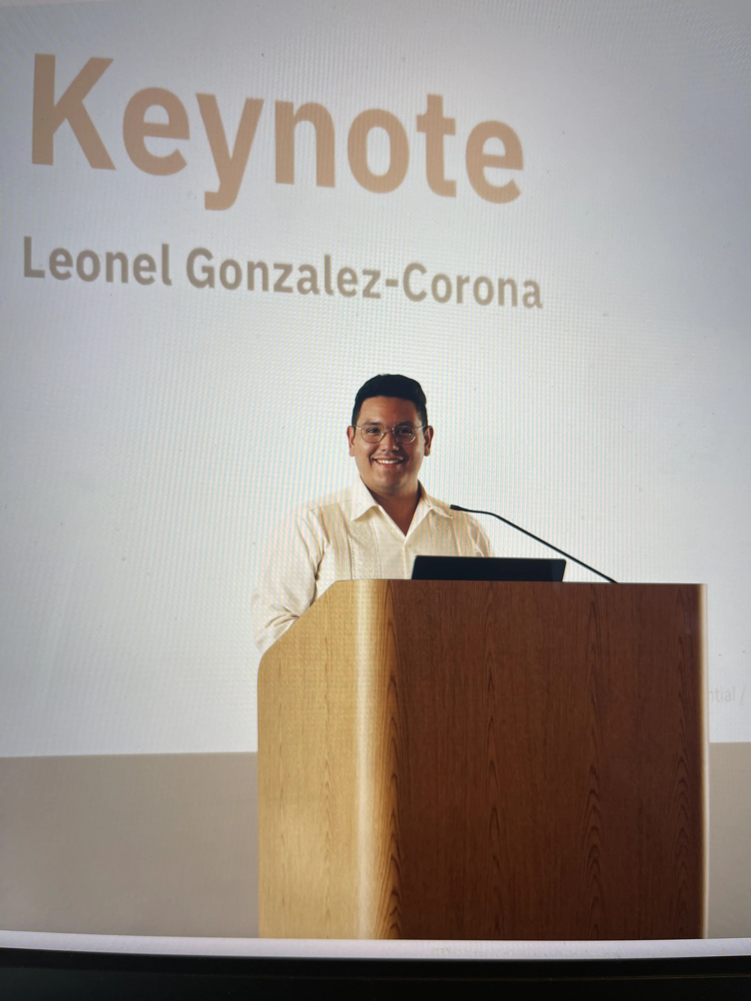 A man standing at a wooden podium, smiling, with a large presentation screen behind him displaying the words 'Keynote' and 'Leonel Gonzalez-Corona'.
