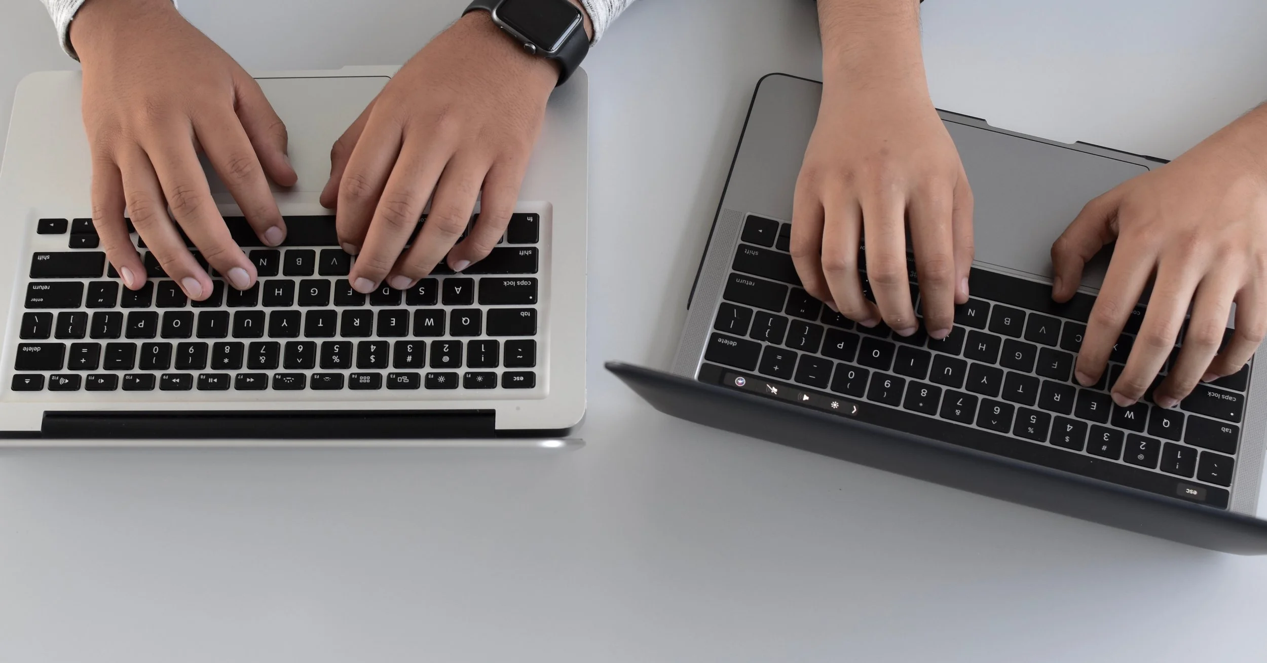 Two people working on laptops, one with a silver MacBook and the other with a gray one, on a light-colored table. Both are typing on their keyboards, with the person on the left wearing a smartwatch.
