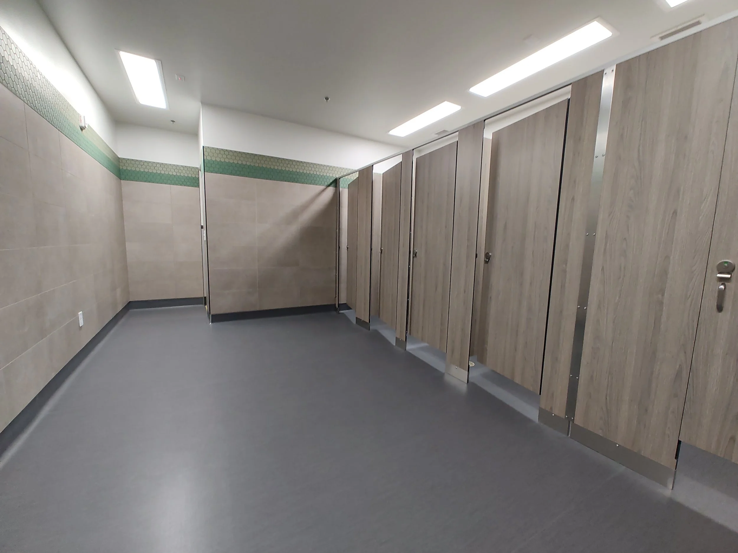 Empty public restroom with stalls, wood doors, tiled walls, and gray flooring.