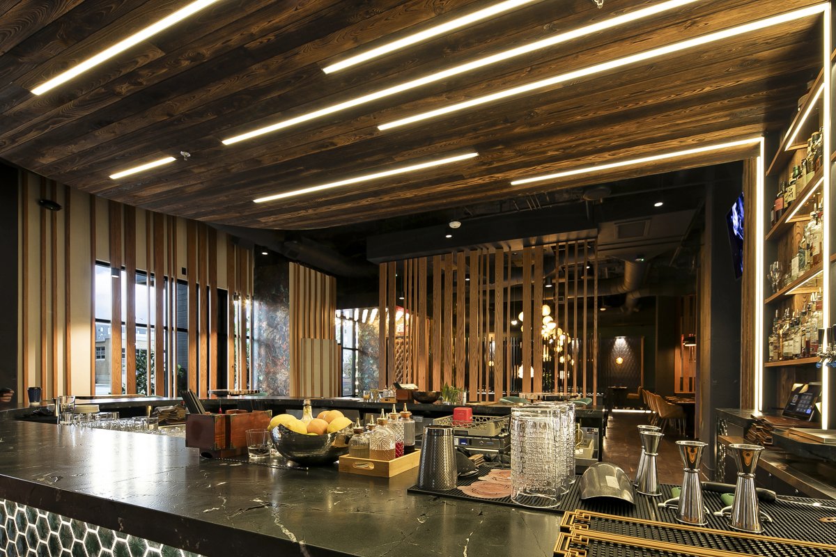 Inside a modern bar with dark wood accents, a black marble counter, and glassware and bottles on display. The ceiling has wooden panels with integrated lighting.