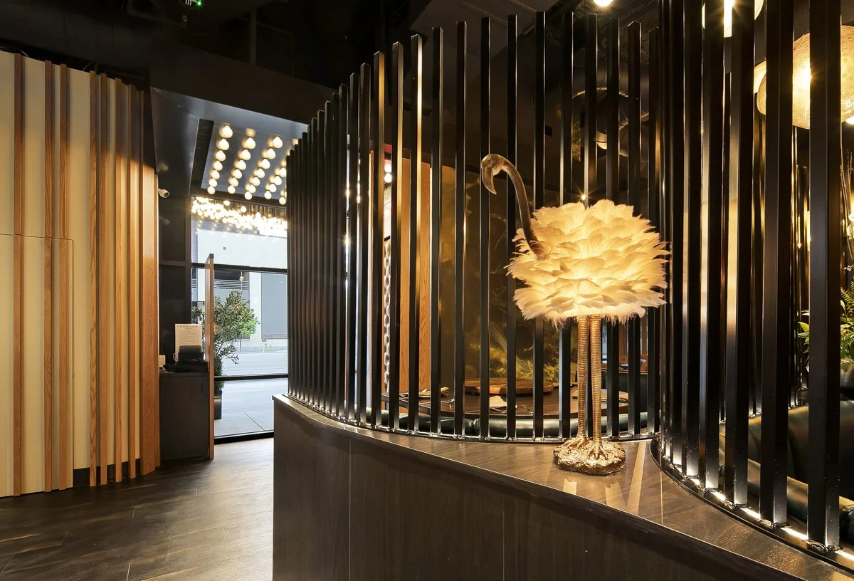 Decorative desk lamp with a flamingo shape and a feathered shade on a dark wood counter in a modern hotel lobby with black and wood accents.