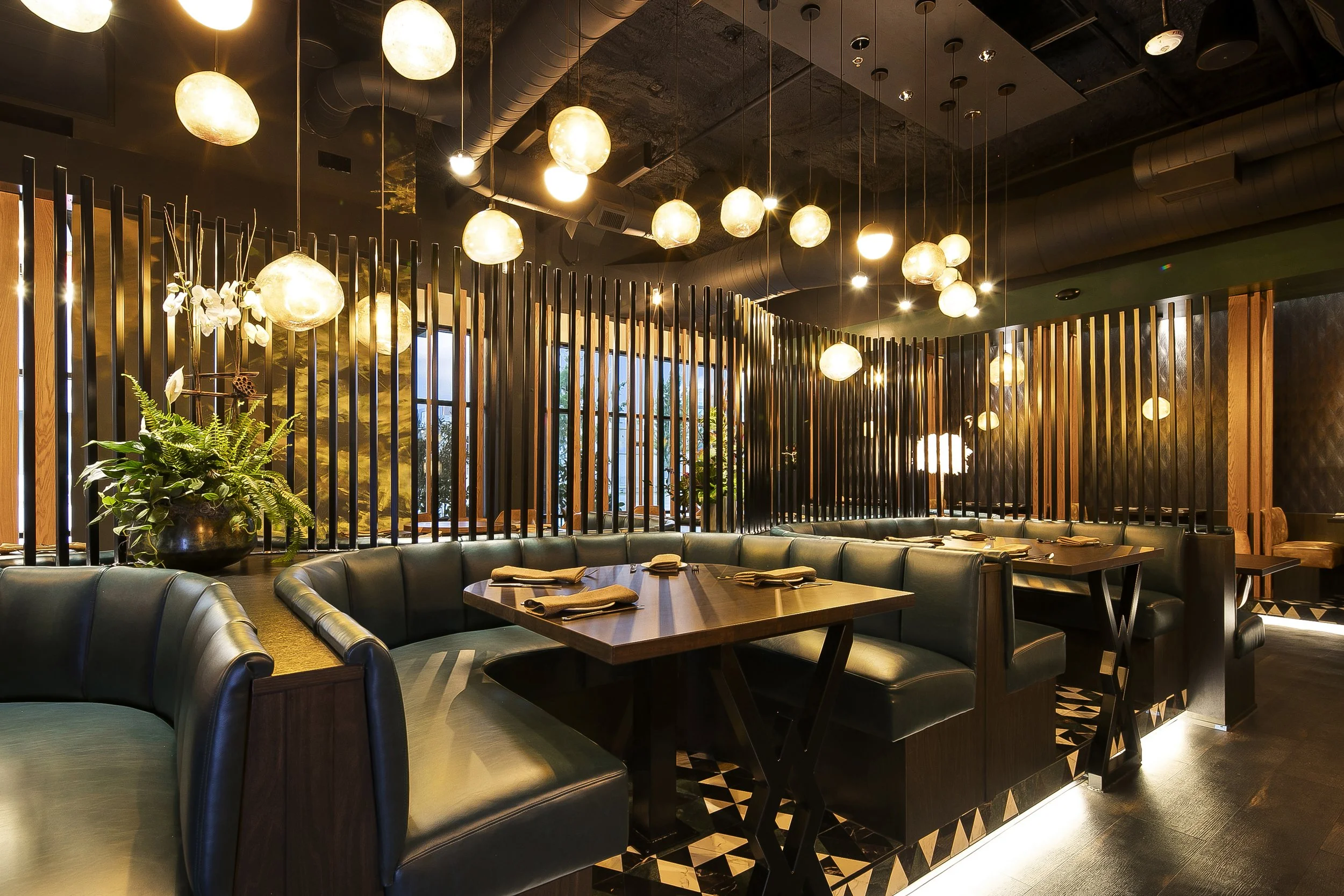Interior of a modern restaurant with a black and brown color scheme, round pendant lights hanging from the ceiling, leather booth seating, and a decorative wooden partition with plants and large windows in the background.