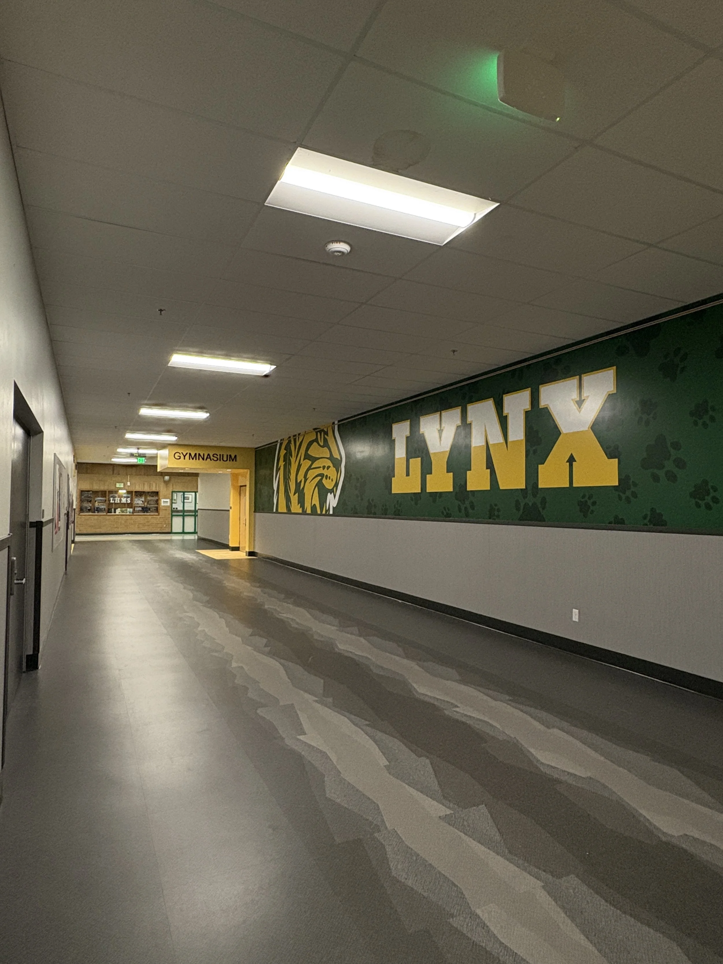 School hallway with a large green wall sign displaying the word "Lynx" and a lynx mascot, with a yellow sign at the end indicating the gymnasium.