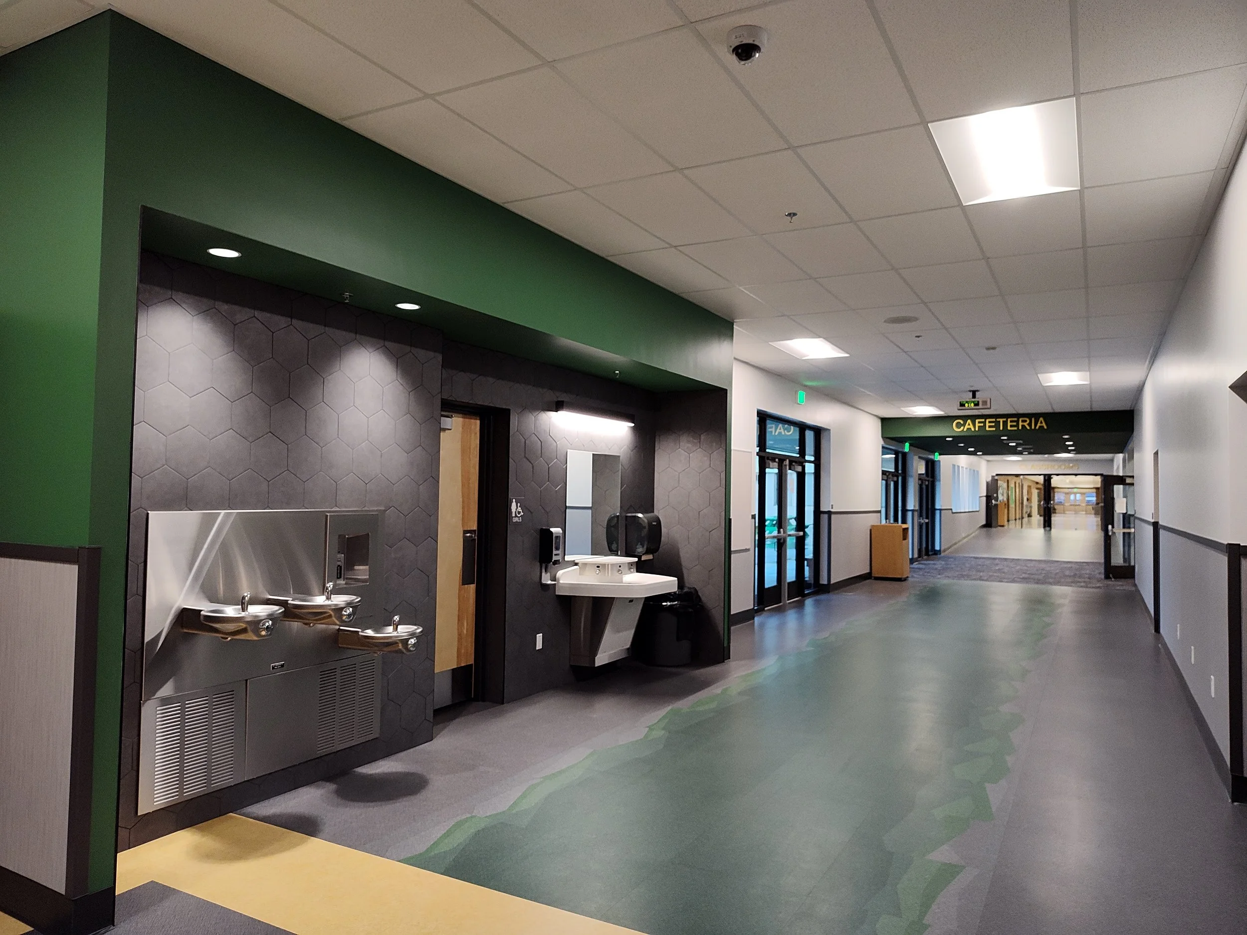 An indoor hallway in a school or office building with a water fountain, a sink, and a sign indicating a cafeteria at the end of the corridor.