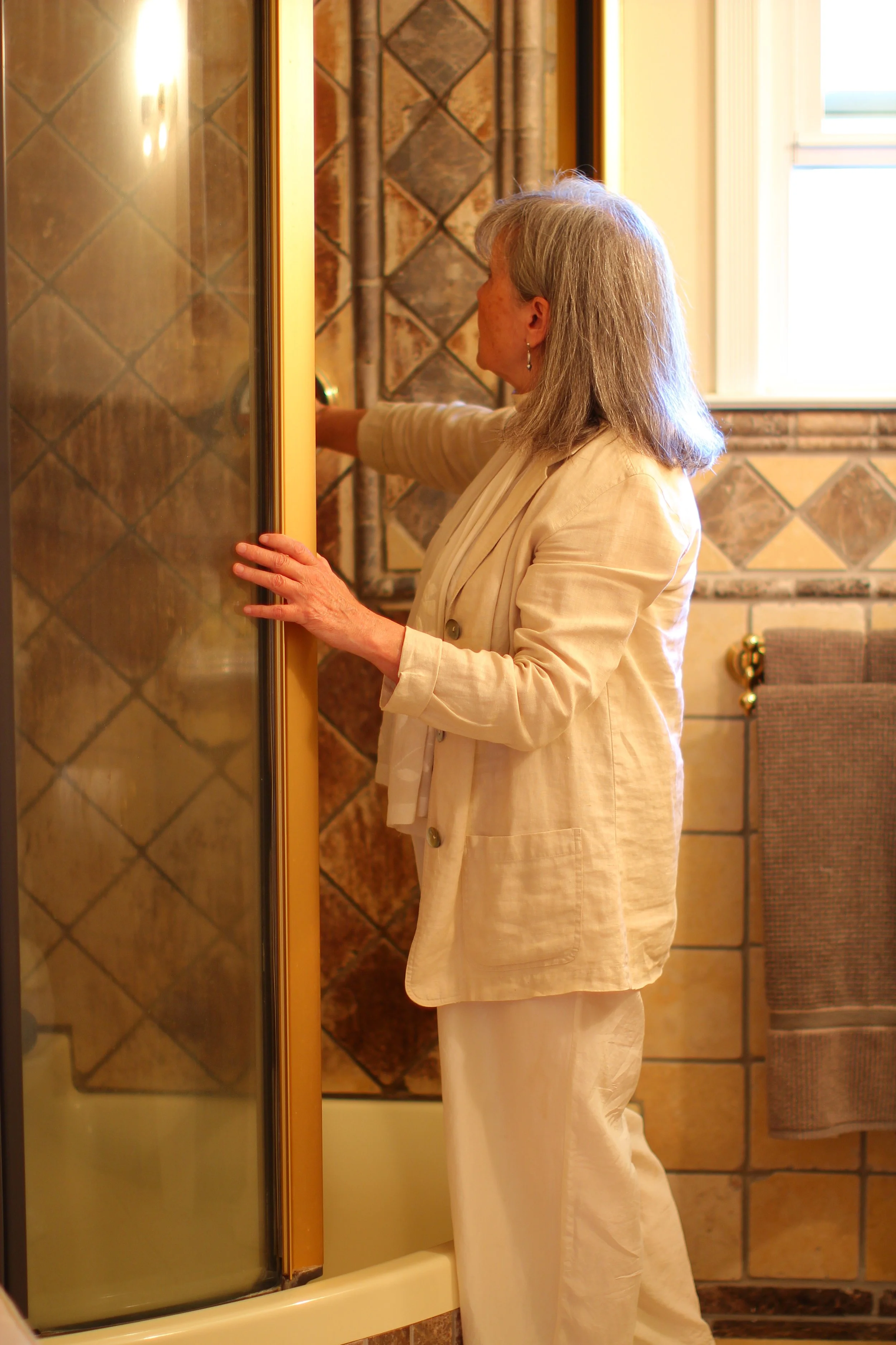 A woman with gray hair wearing a cream-colored suit is adjusting a shower door in a bathroom with tiled walls and a window.