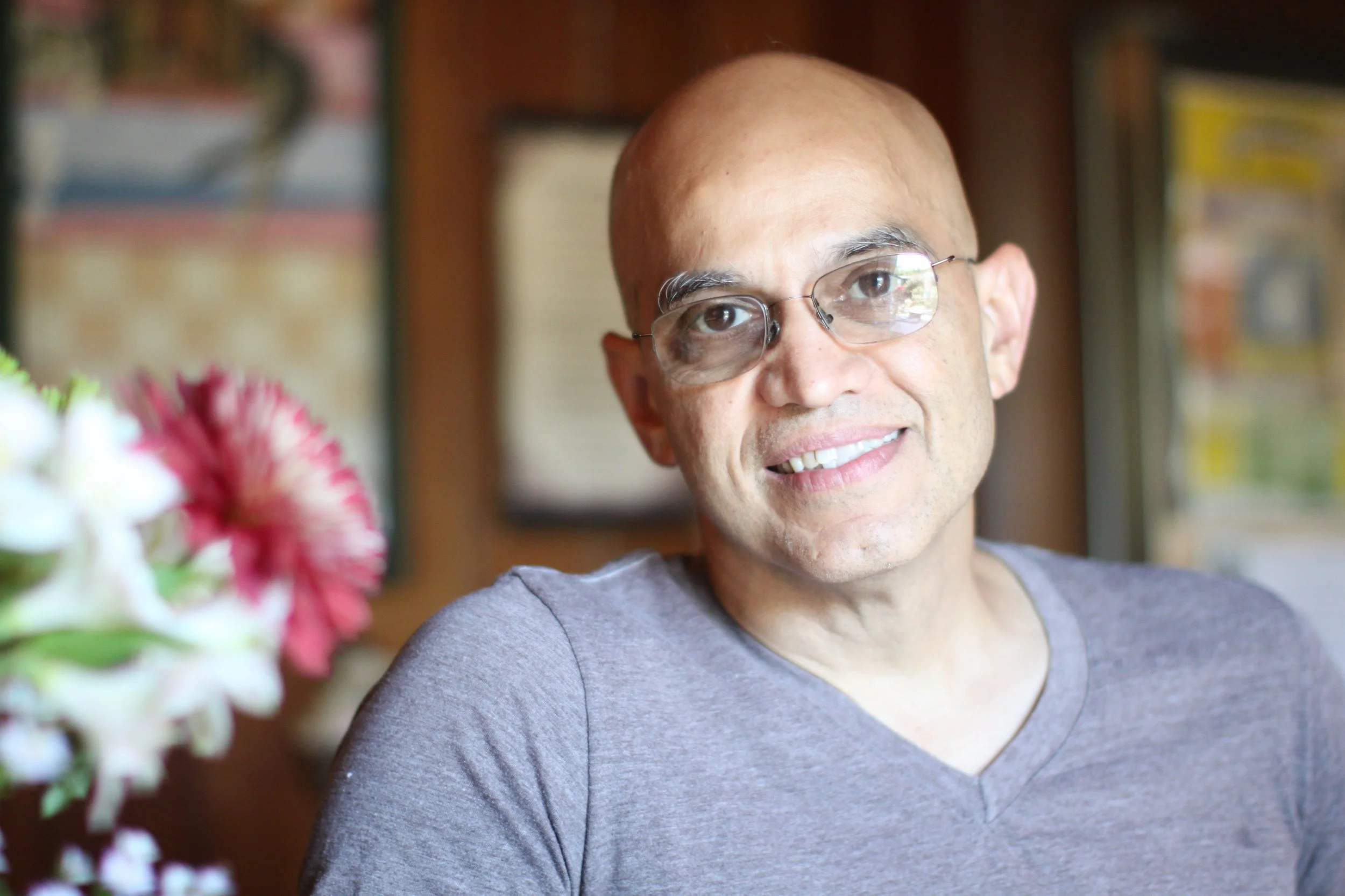 A man with a shaved head and glasses smiling indoors with flowers and framed pictures in the background.
