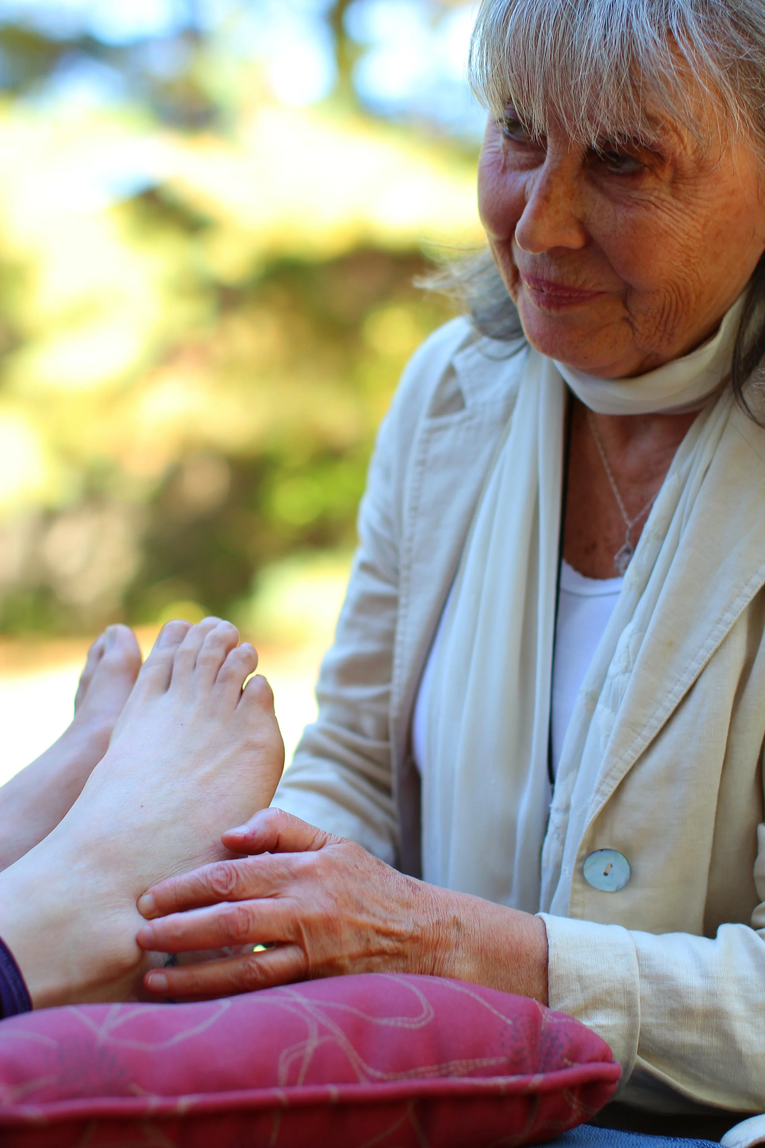 An elderly woman with gray hair and a white jacket gently holds the hand of a younger person, with both hands resting on a pink cushion outdoors with blurred green and yellow foliage in the background.