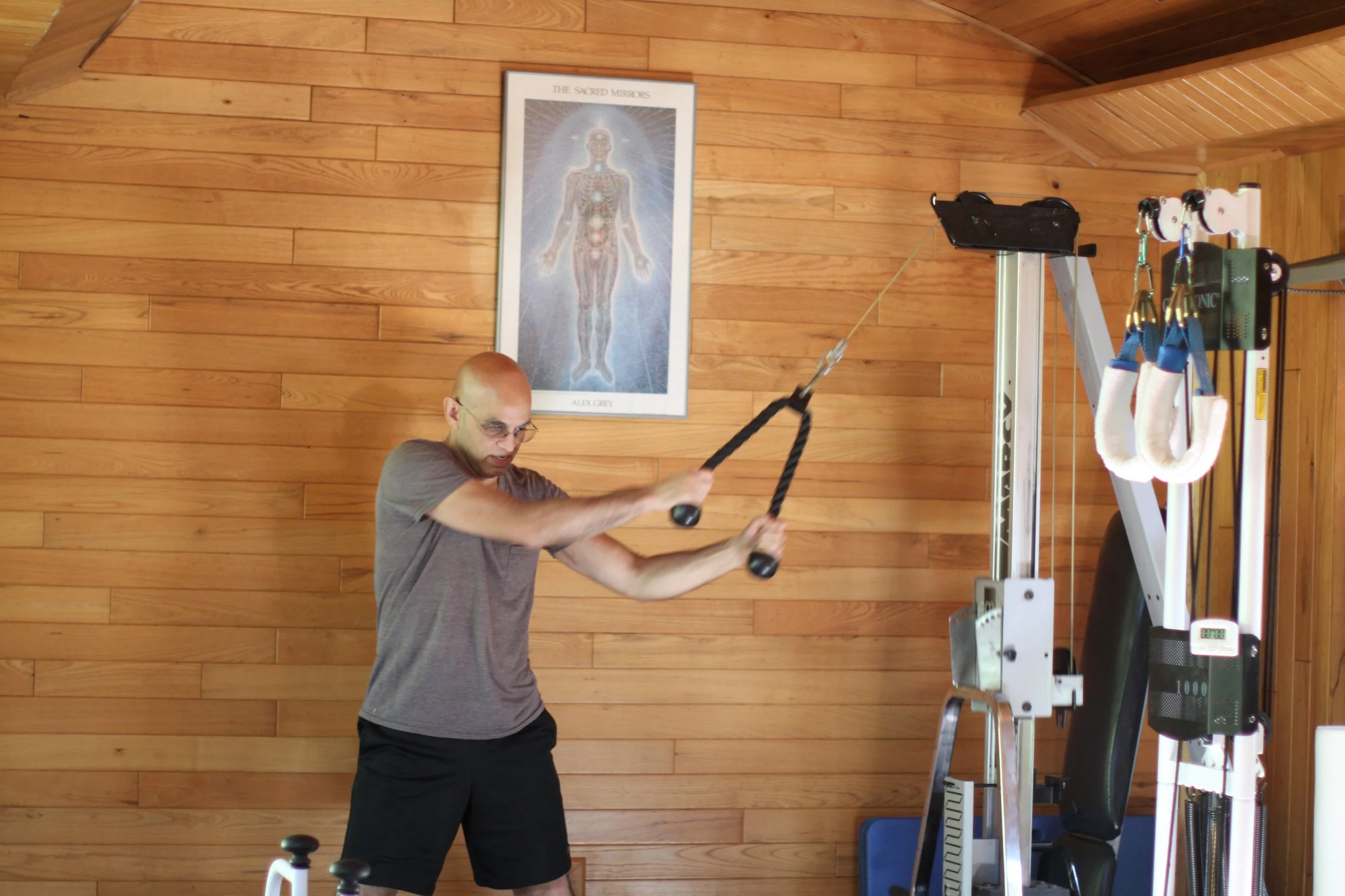 Man using a cable machine for strength training in a wooden-paneled gym, with a framed anatomical chart on the wall.