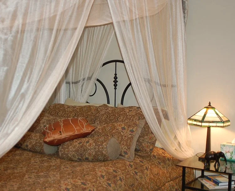 A bed with a decorative iron headboard, surrounded by sheer curtains. The bedding and pillows have a paisley and floral pattern, including a pillow in the shape of a piece of meat. There is a side table with a stained glass lamp, a small elephant fig