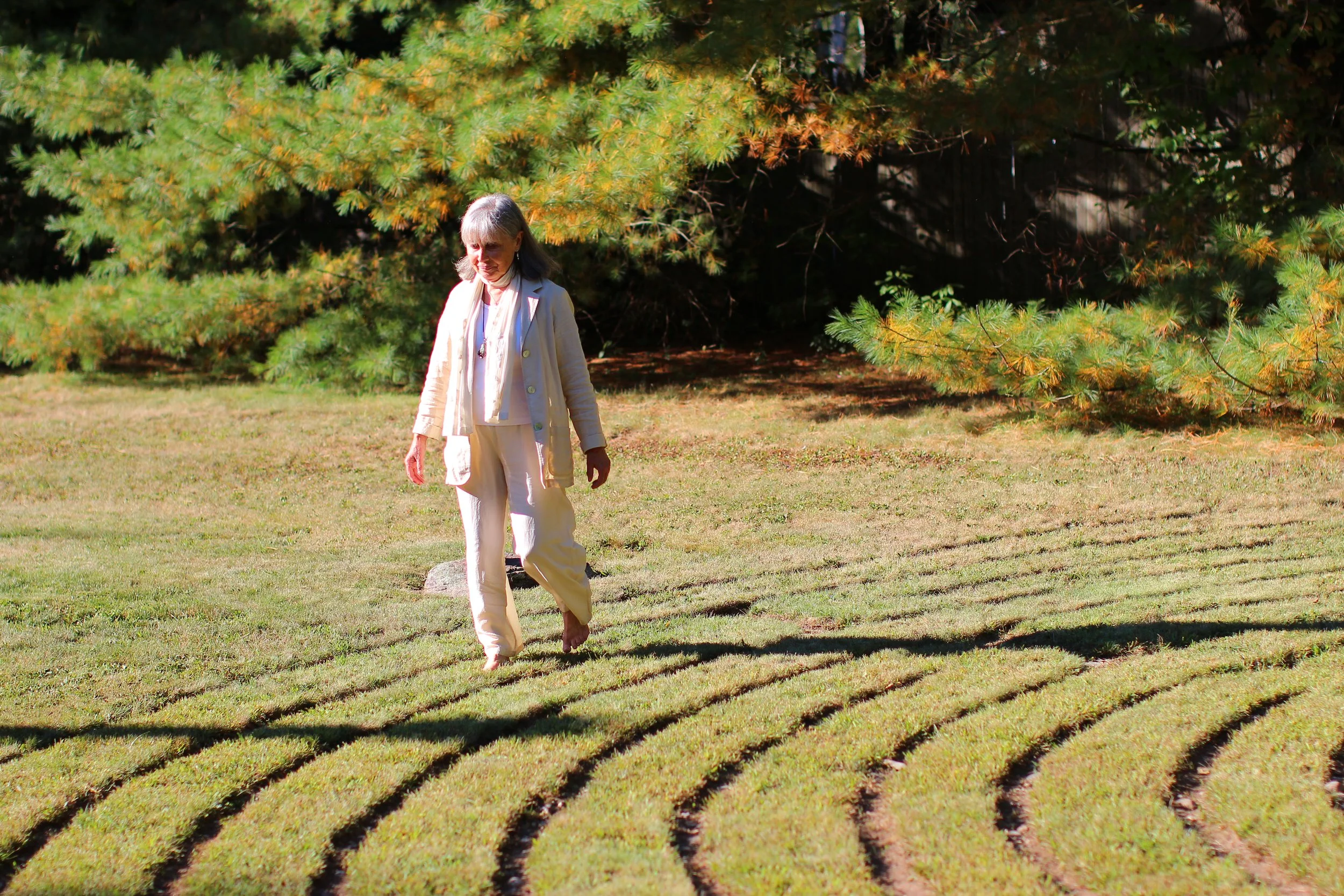 An older woman with gray hair walking bare feet through a grassy area with curved rows of recently planted grass. She is wearing a white jacket and light-colored pants, with a necklace. In the background, there are green trees with some yellowish lea