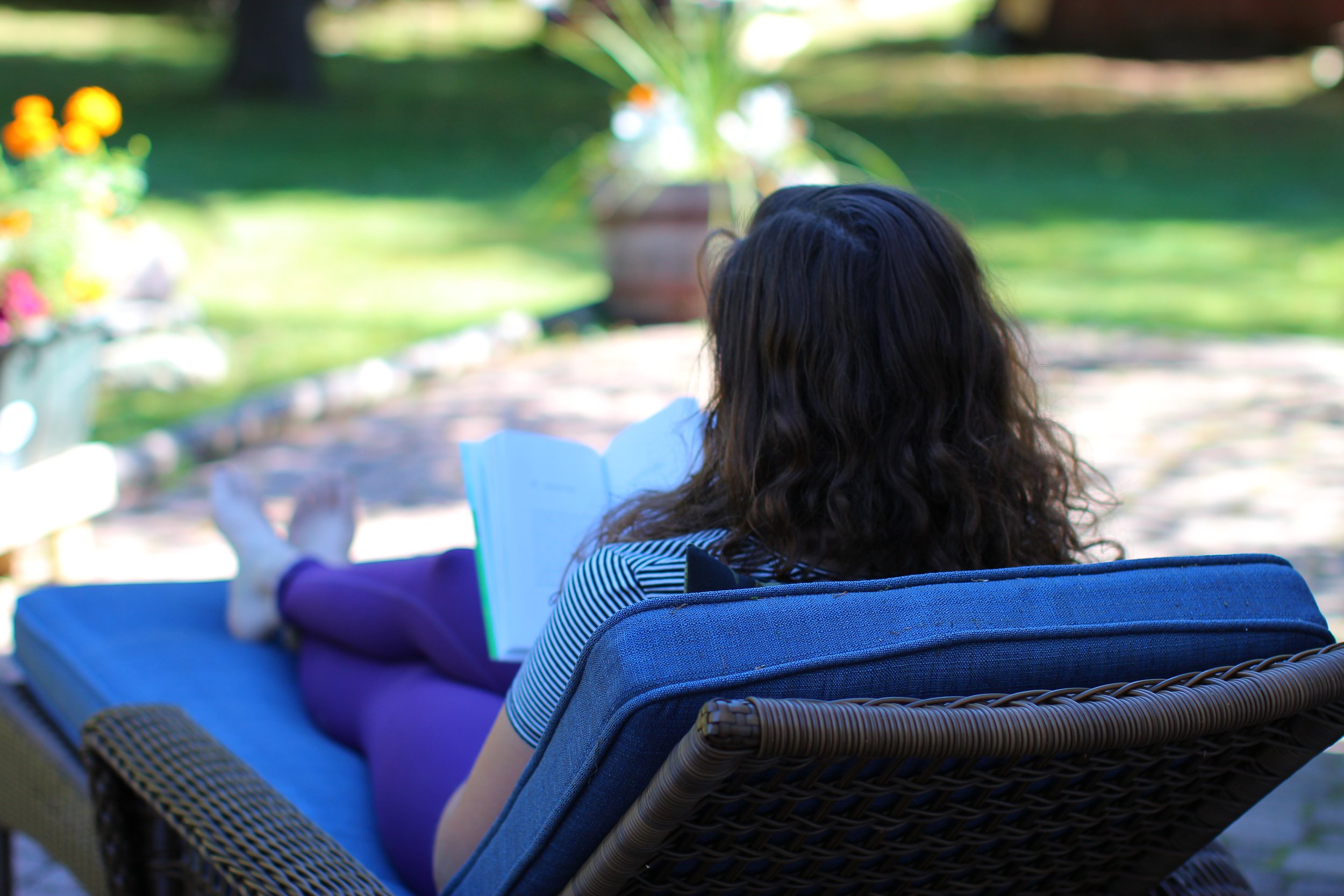 A woman with curly hair is sitting on a wicker lounge chair outdoors, reading a book in a garden with greenery and flowers.
