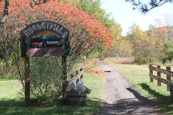 A trail entrance sign for Hurlburtville with the text "Rails to Trails," surrounded by colorful fall foliage, along a dirt trail with a wooden fence on one side.