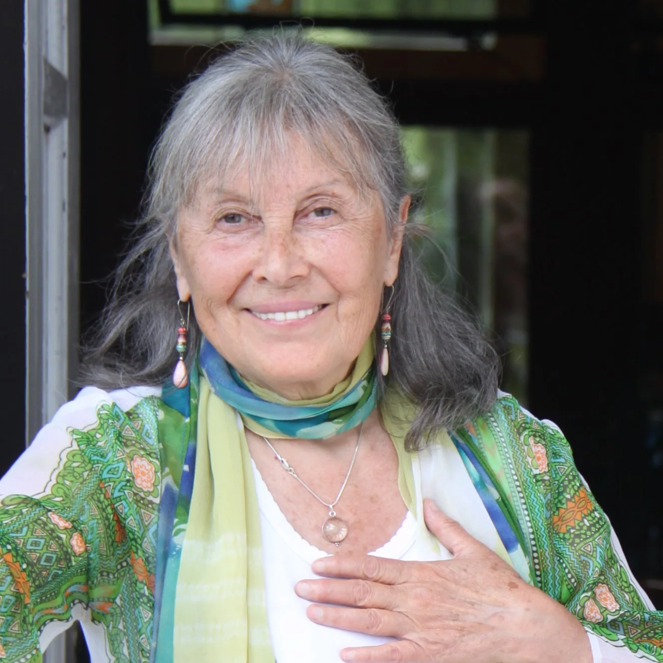 Older woman with gray hair smiling and touching her chest, wearing colorful earrings, a necklace with a round pendant, a white top, and a multicolored scarf, outdoors with blurred greenery in the background.