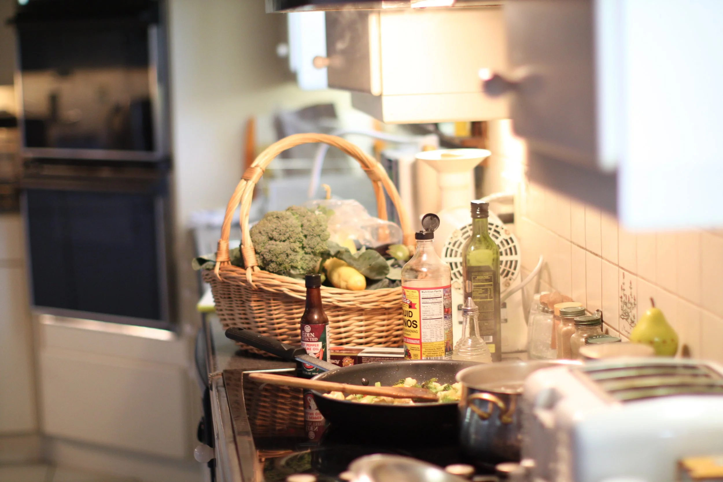 Kitchen countertop with a basket of vegetables, a stove with a frying pan, bottles of cooking oils and condiments, and various kitchen items in a cozy home kitchen.