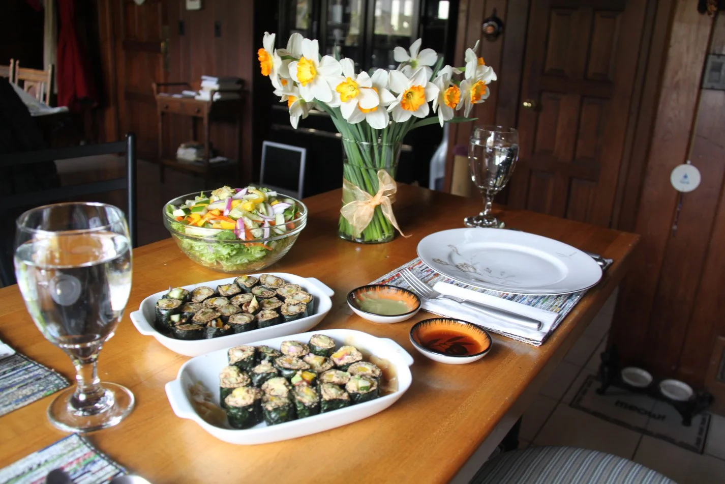 A wooden dining table set with a large vase of daffodils, a bowl of salad, two dishes of sushi rolls, a plate with a decorative edge, two small soy sauce dishes, a glass of water, and a glass of wine in a cozy dining room.