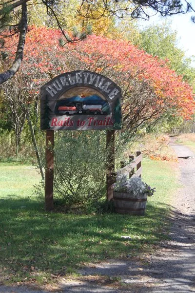 HURLEYVILLE sign with a vintage truck illustration and the phrase "Rails to Trails" beneath it, surrounded by trees with colorful fall foliage and a gravel path.