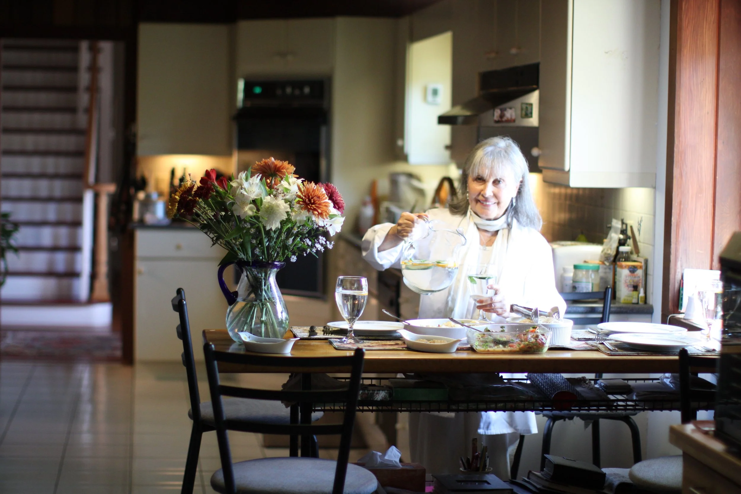 An elderly woman with gray hair, smiling, pours water from a glass pitcher into a small glass at a dining table set with a floral centerpiece, salad, and glass of water, in a bright kitchen.