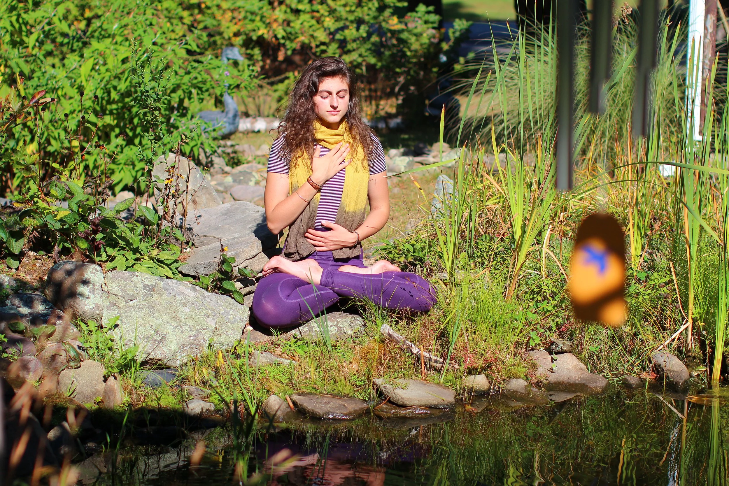 A woman practicing yoga or meditation outdoors near a small pond surrounded by rocks and green vegetation, with sunlight streaming through trees.