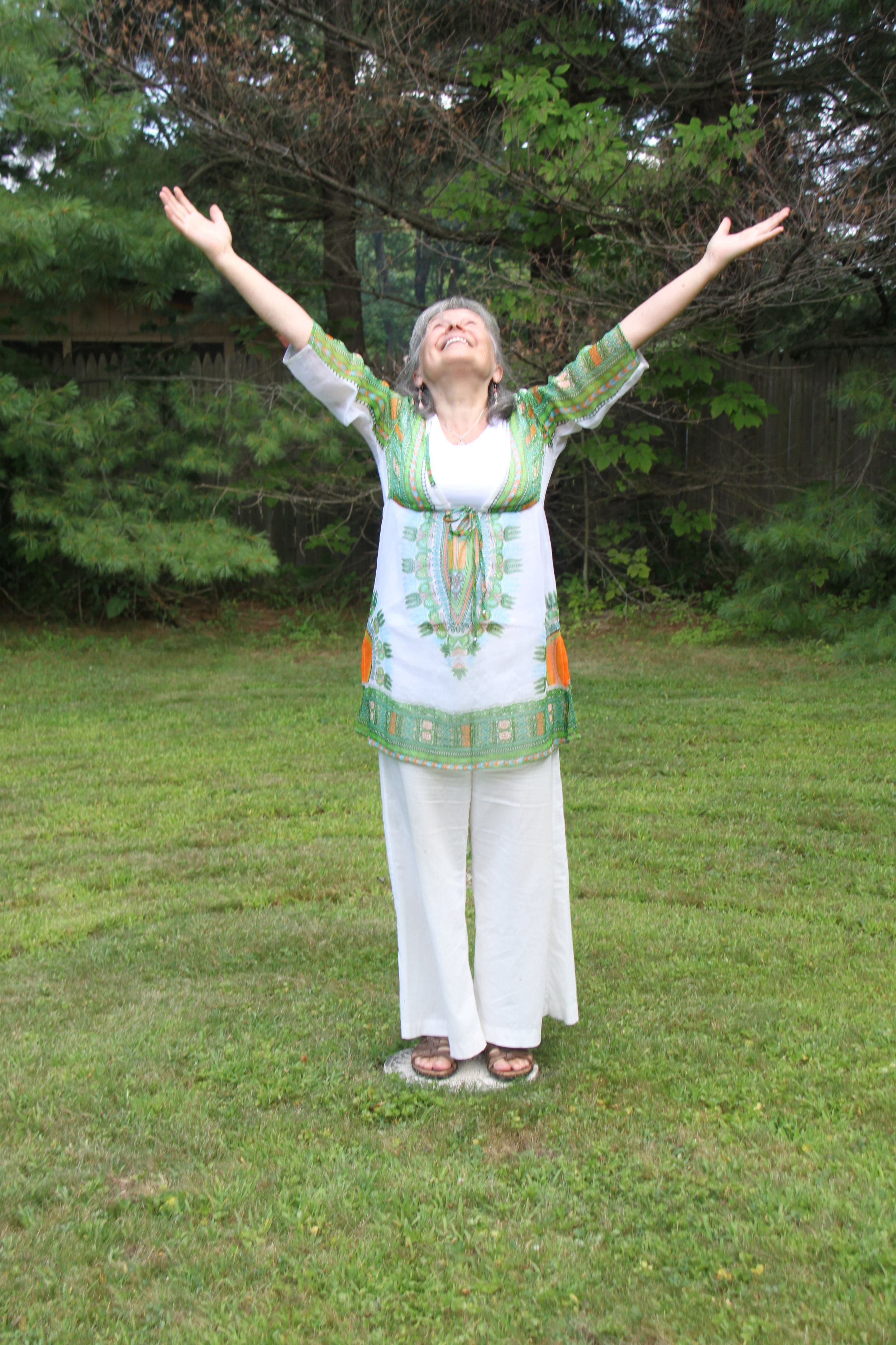 A woman dressed in colorful, bohemian clothing stands outdoors on grass with her arms raised and head tilted back, appearing joyful and liberated.