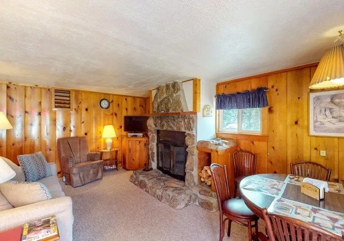 Cozy living room with wood-paneled walls, a stone fireplace, a beige sofa with pillows, an armchair, a small side table with a lamp, a mounted TV, a round dining table with chairs, and a window with a blue valance.