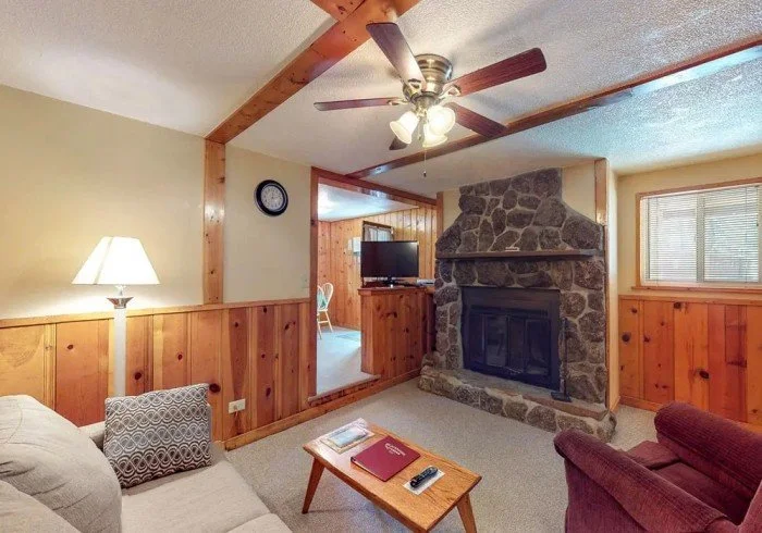 Cozy living room with wood-paneled walls, fireplace with a stone surround, ceiling fan with lights, sofa, armchair, and a small wooden coffee table with books and a TV in an adjacent room.