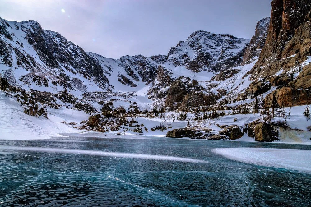 Snow-covered mountains around a partially frozen lake under a cloudy sky.