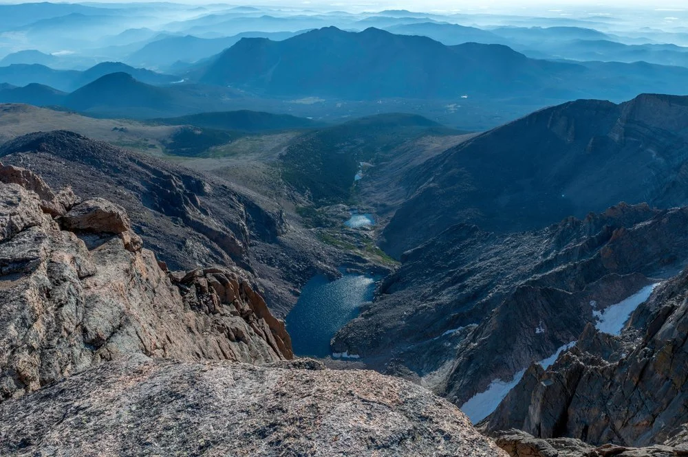 Aerial view of mountain landscape with rugged rocks, a lake, and patches of snow, with distant mountain ranges under a blue sky.