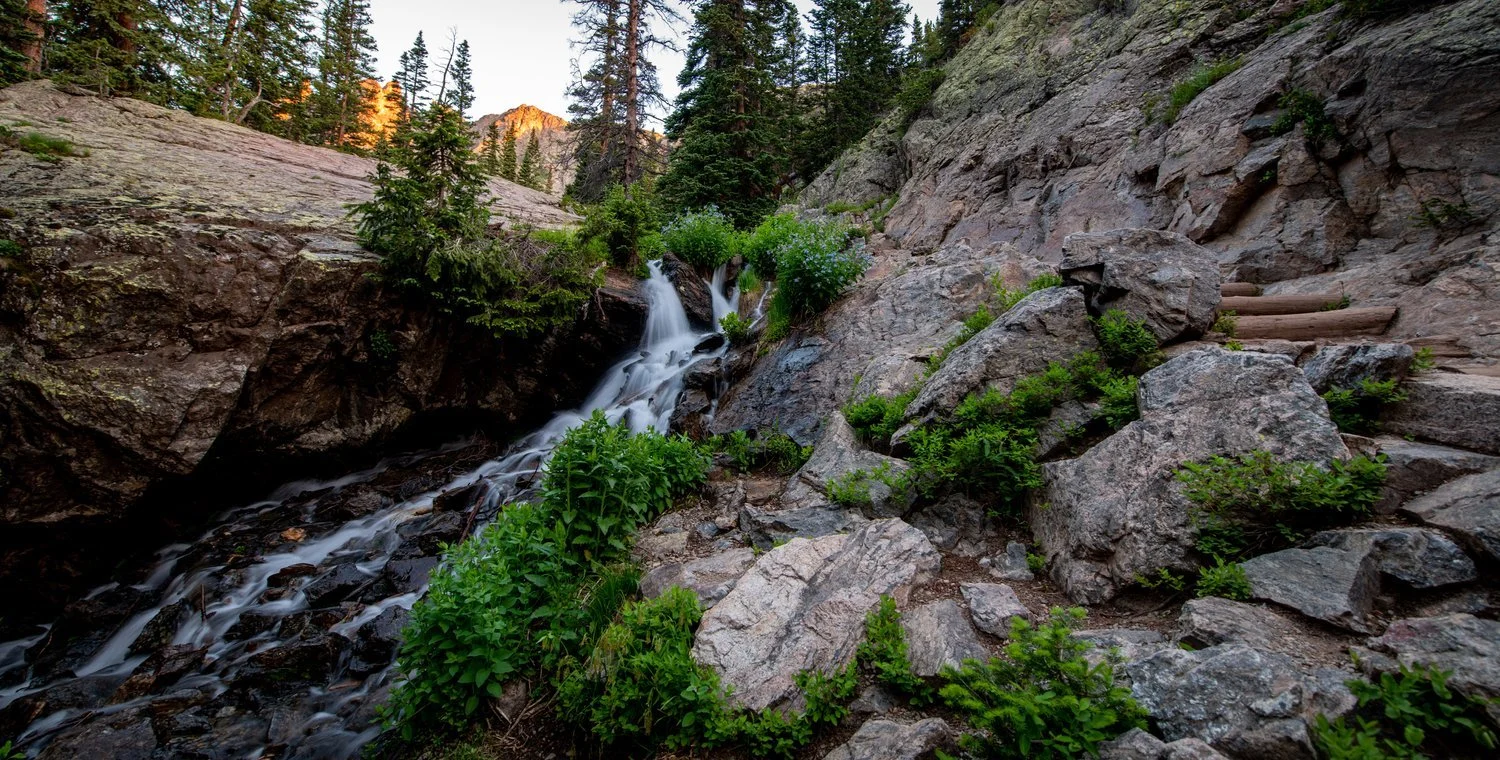 A rocky mountain landscape with a small waterfall flowing down the rocks surrounded by green shrubs and pine trees.