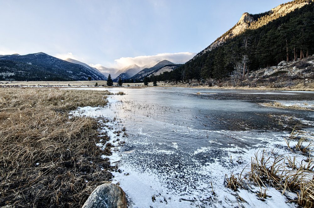 A winter landscape with snow-covered grass and a partially frozen river, surrounded by mountains and trees under a partly cloudy sky.