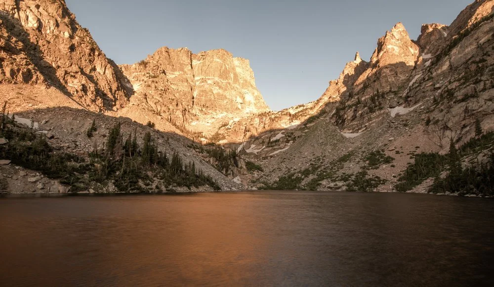 A scenic mountain landscape with a lake in the foreground and rugged cliffs illuminated by sunlight in the background.