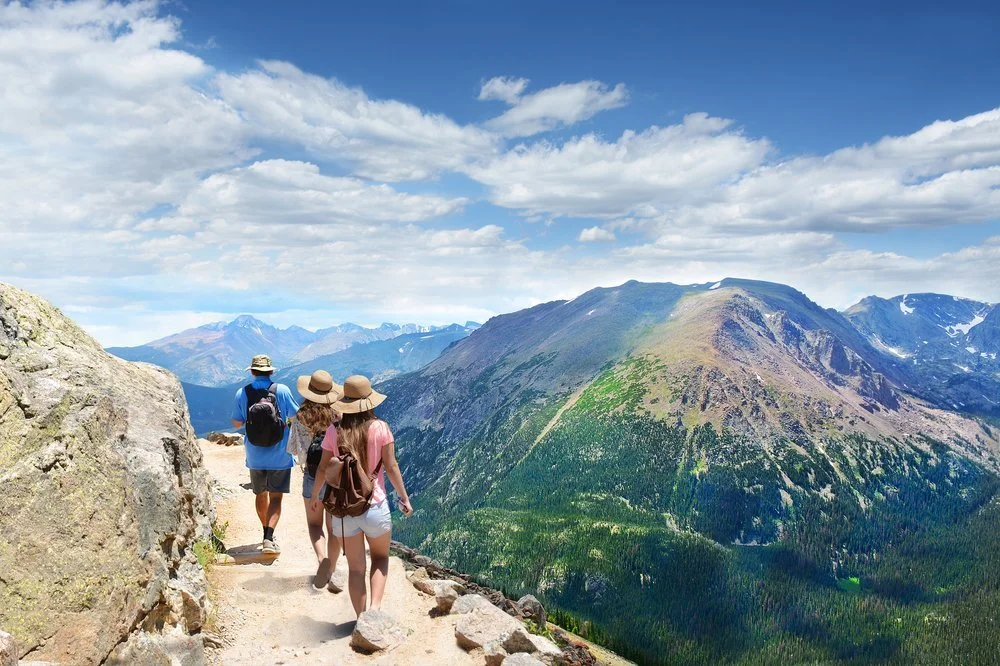 Four people hiking on a mountain trail with a mountain range and blue sky with clouds in the background.