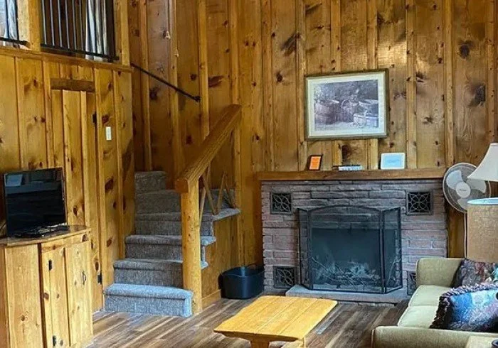 Living room with wood-paneled walls, a brick fireplace, a yellow couch, a small wooden table, and a staircase with carpeted steps.
