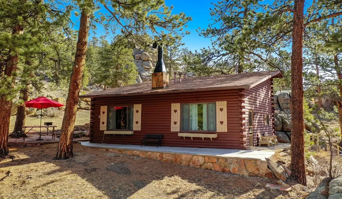 A cozy log cabin in a wooded area with tall pine trees, a stone foundation, and a chimney, featuring shutters with heart shapes, a large window, a picnic table with a red umbrella, and an outdoor bench, under a clear blue sky.