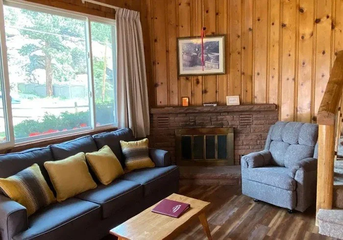 Living room with wooden walls, large window with curtains, black sofa with beige and patterned pillows, gray armchair, fireplace with brick surround, framed picture of a flamingo above the fireplace, and wooden flooring.