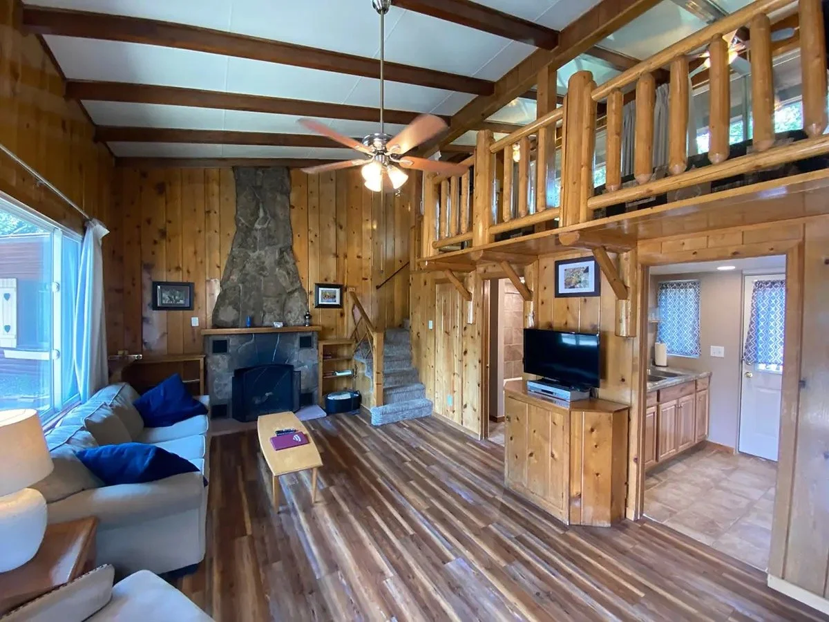 Interior of a log cabin living room with wood panel walls, wooden ceiling beams, a stone fireplace, a ceiling fan, couches, a small coffee table, a television, and a kitchen area with wooden cabinets.